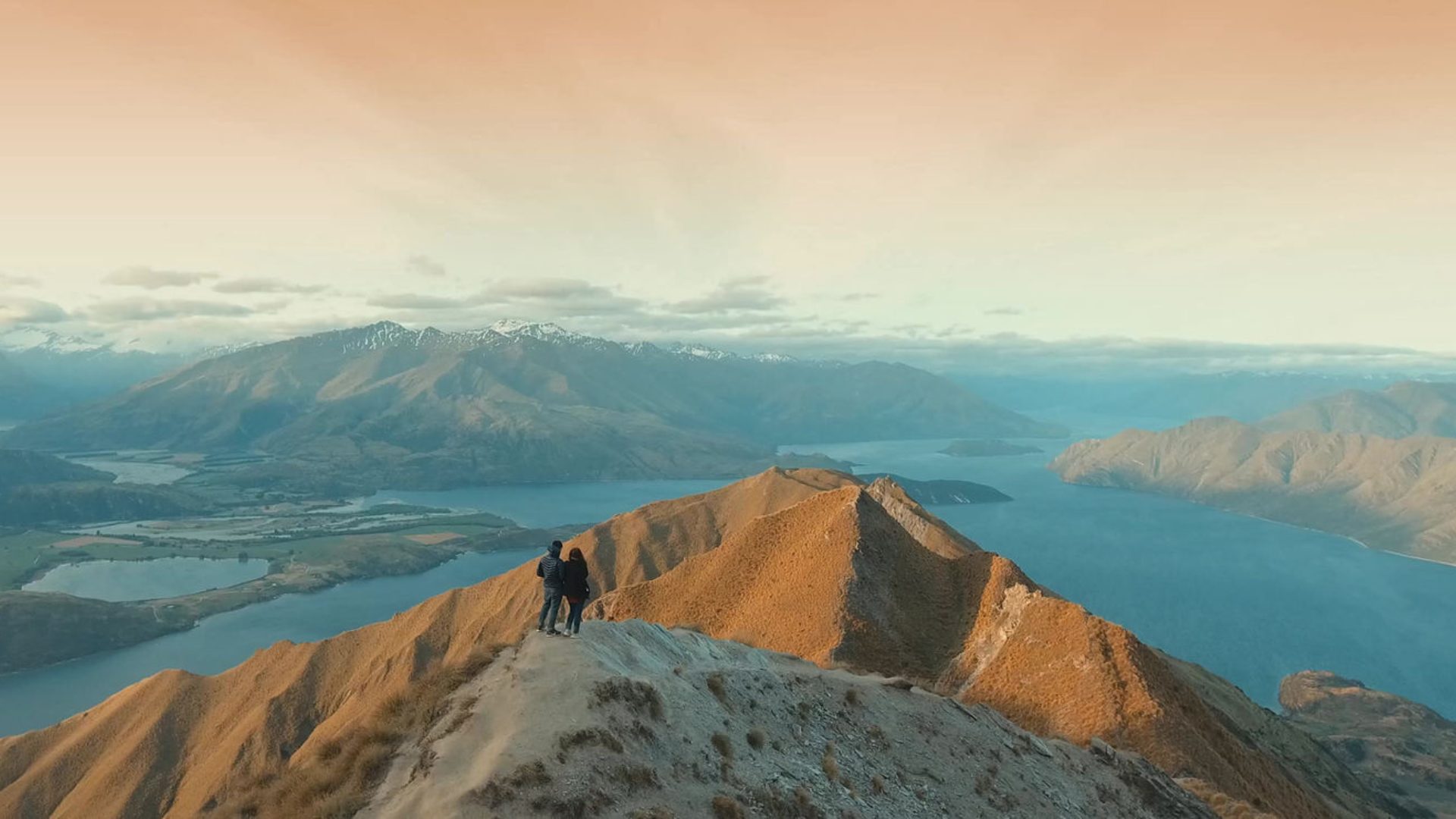 Two people standing on a mountain ridge above a lake